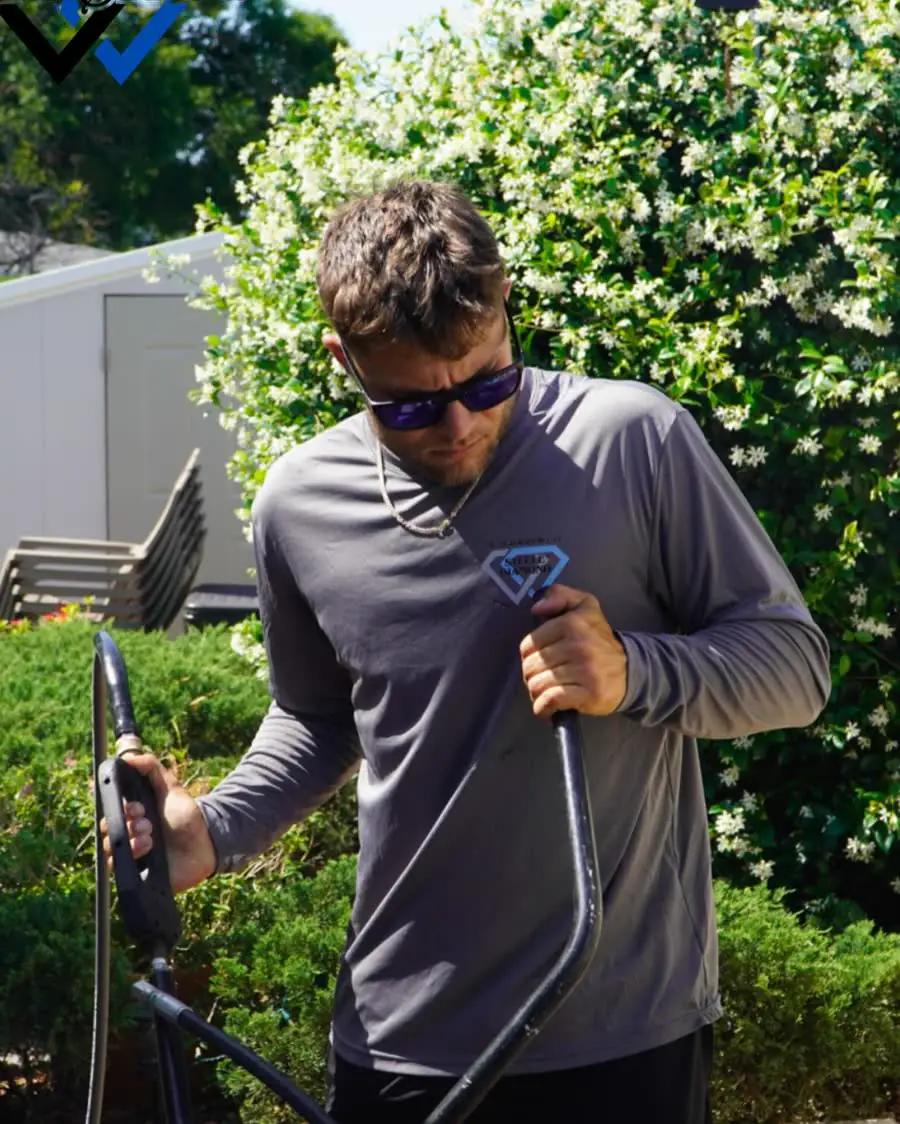 One of the Steele Diamond crew in sunglasses and a long-sleeve work shirt, holding a pressure washer wand among green shrubs.
