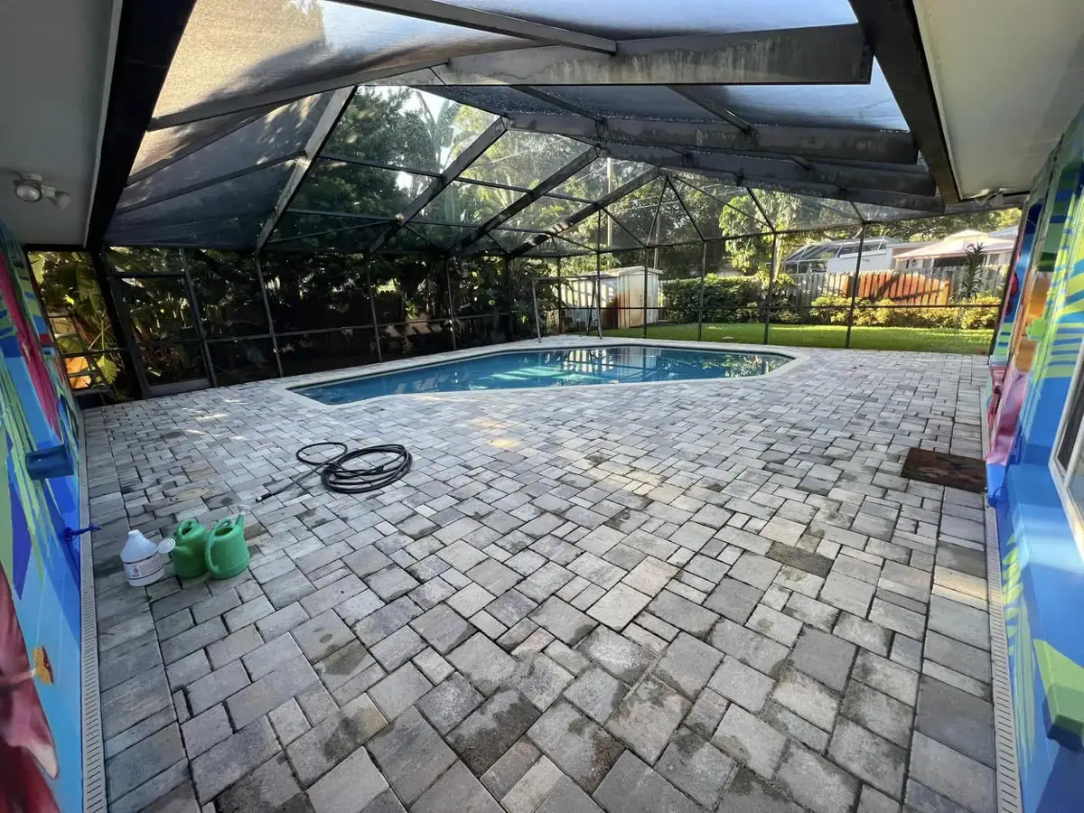 Before: a paver pool deck under a screen cage, grey and faded, a hose and a bucket on it.