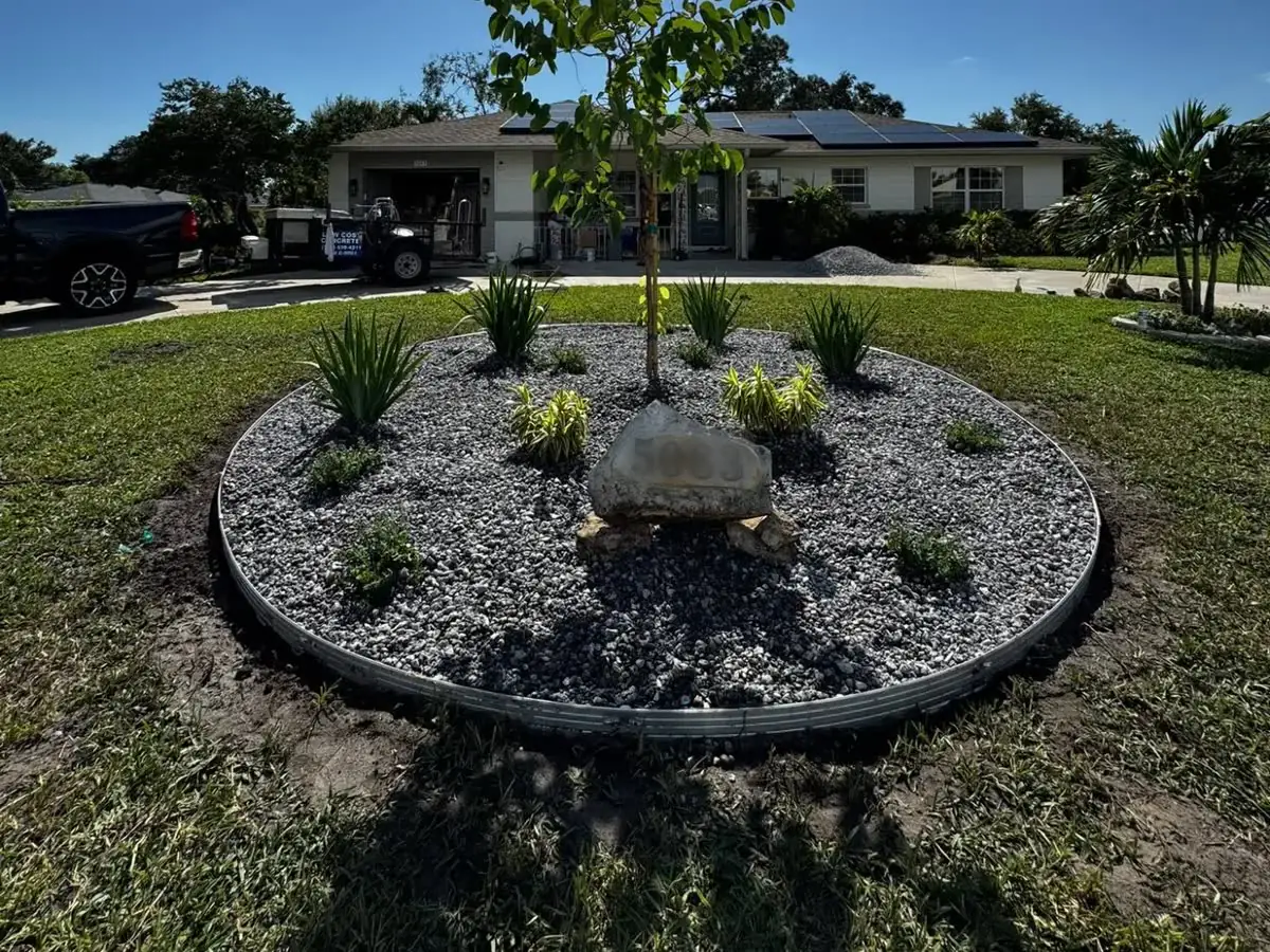 A round front-yard bed with steel edging, grey rock, new plants, a young tree and a boulder.