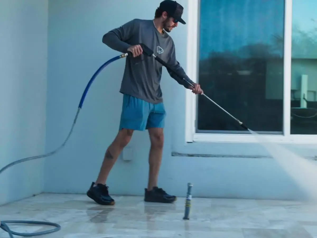 One of the crew pressure washing a travertine pool deck beside sliding glass doors.