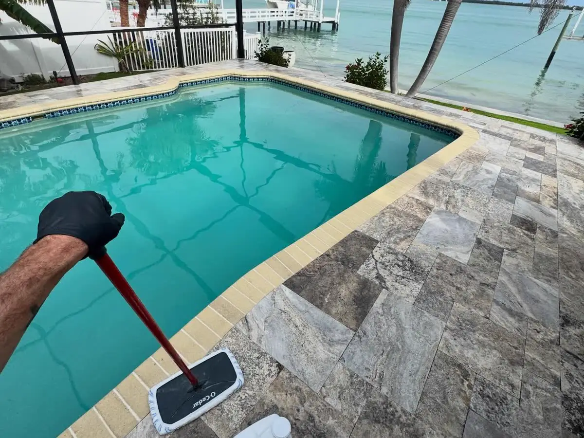 Rolling sealer onto a travertine pool deck inside a screen cage, the water beyond.