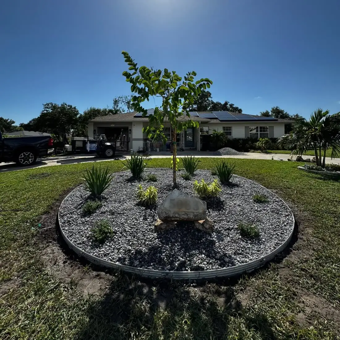 A round front-yard bed with steel edging, grey rock, new plants, a young tree and a boulder.
