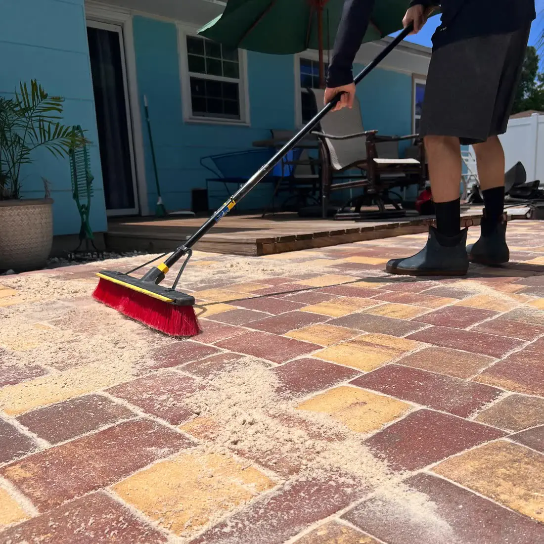 Sweeping joint sand across red, tan and yellow brick pavers with a red push broom, a blue house behind.
