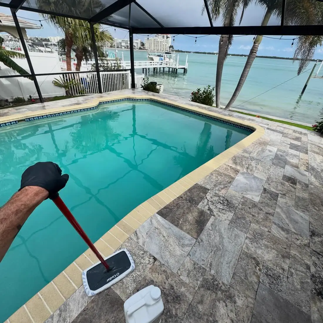 Rolling sealer onto a travertine pool deck inside a screen cage, the water beyond.