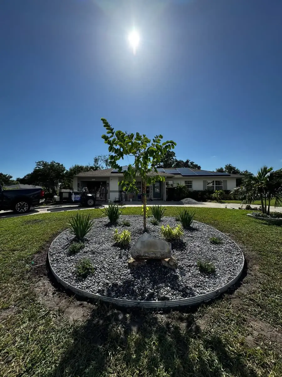 A round front-yard bed with steel edging, grey rock, new plants, a young tree and a boulder.