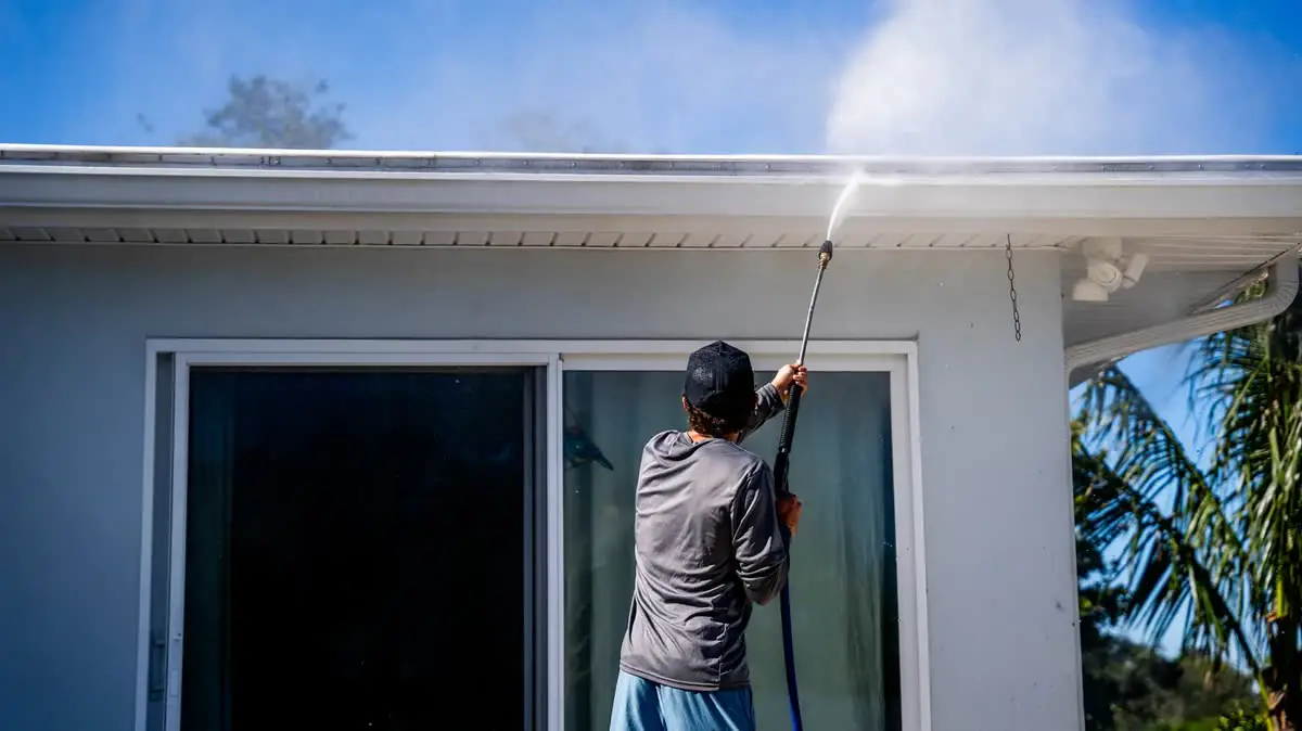 One of the crew washing the eaves of a white house with a wand.