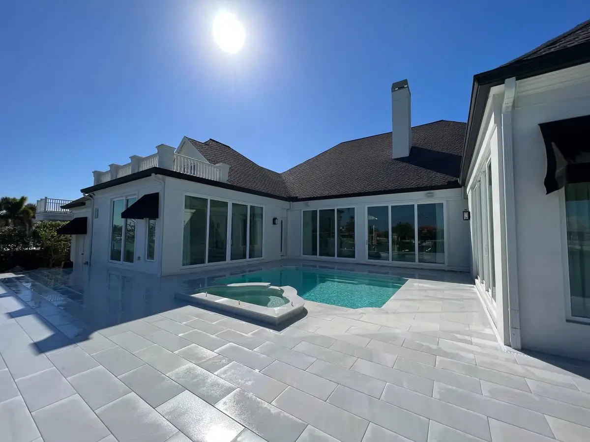 A grey paver pool deck and spa behind a white house.