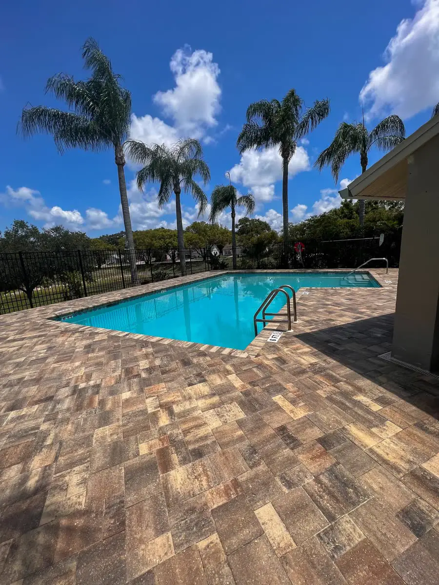A paver pool deck in warm tan and grey pavers around a pool, palms beyond.