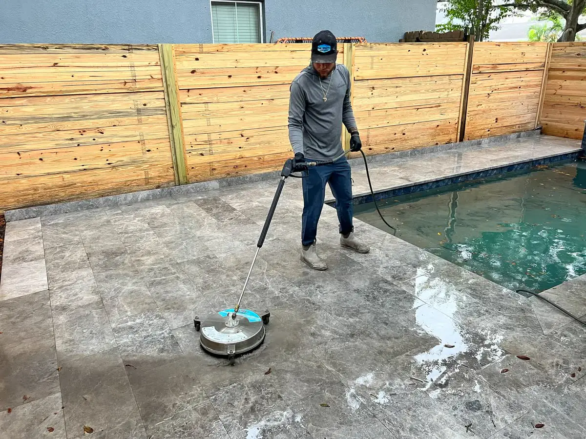 One of the crew with a surface cleaner on a pool deck by a wooden fence.