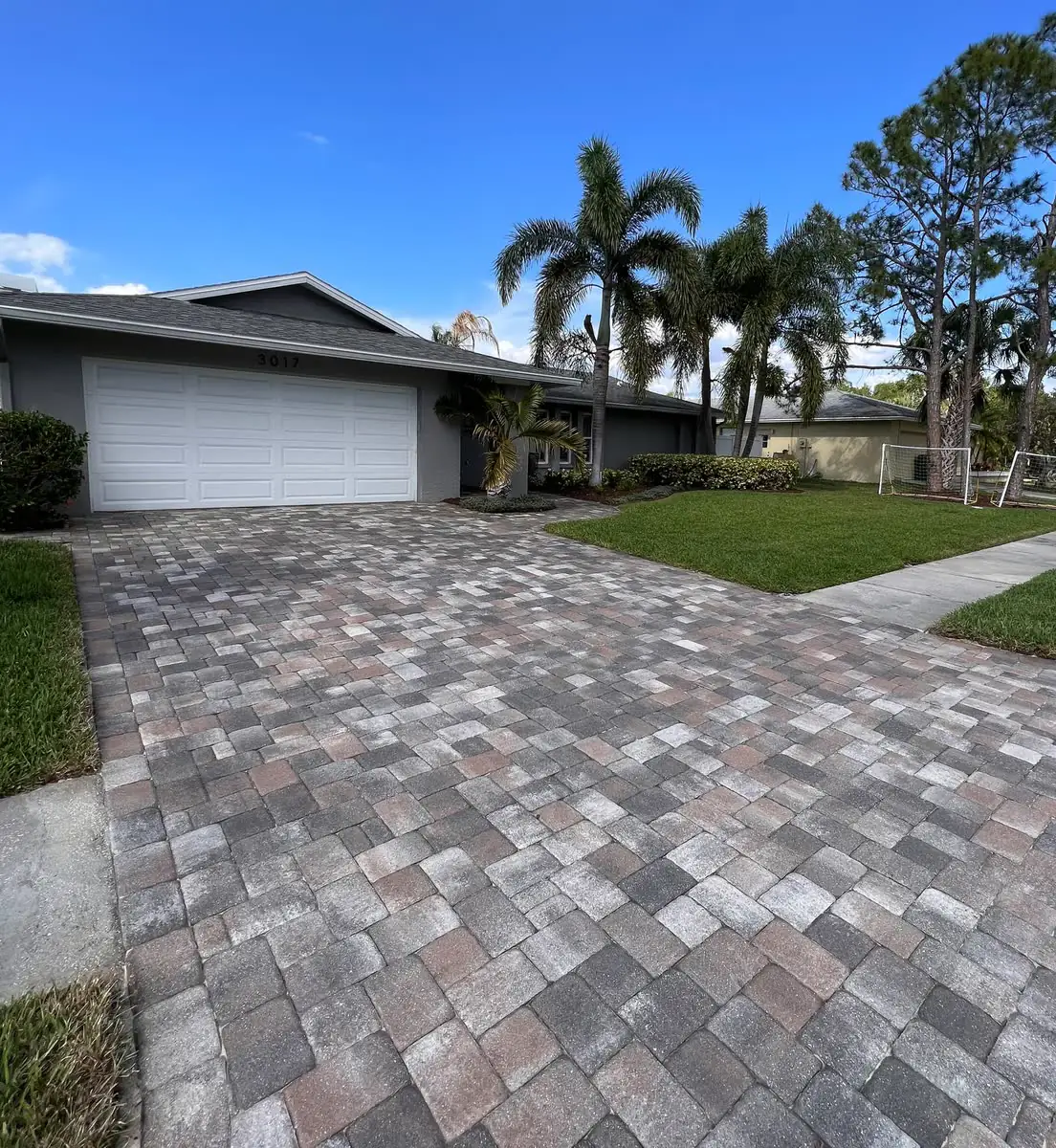 A grey paver driveway in front of a white garage, palms beyond.