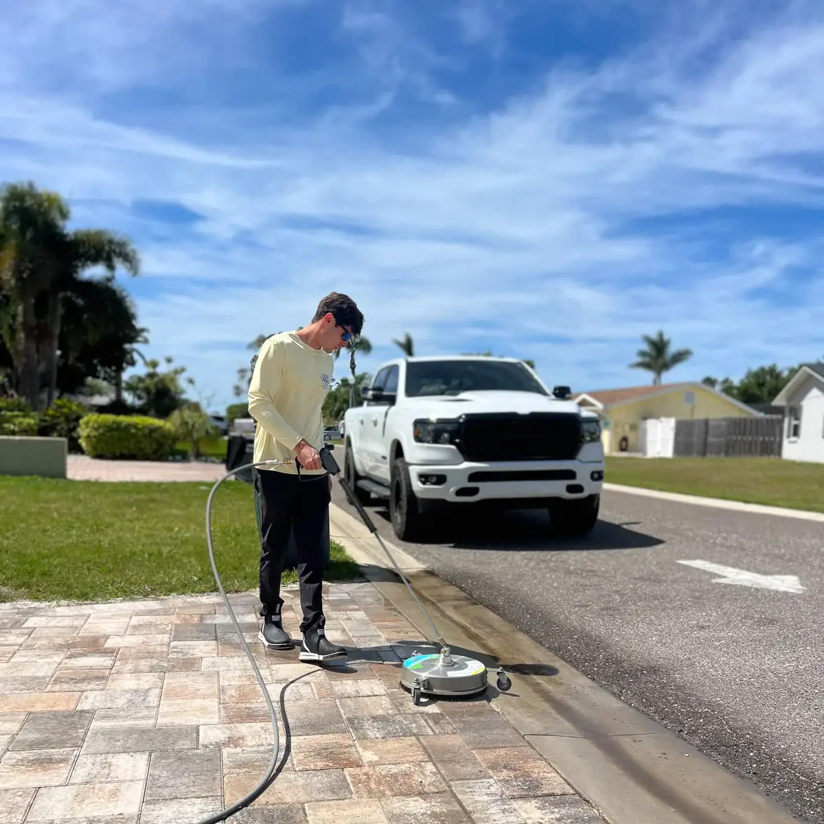 One of the crew running a surface cleaner on a paver driveway, a white pickup behind.