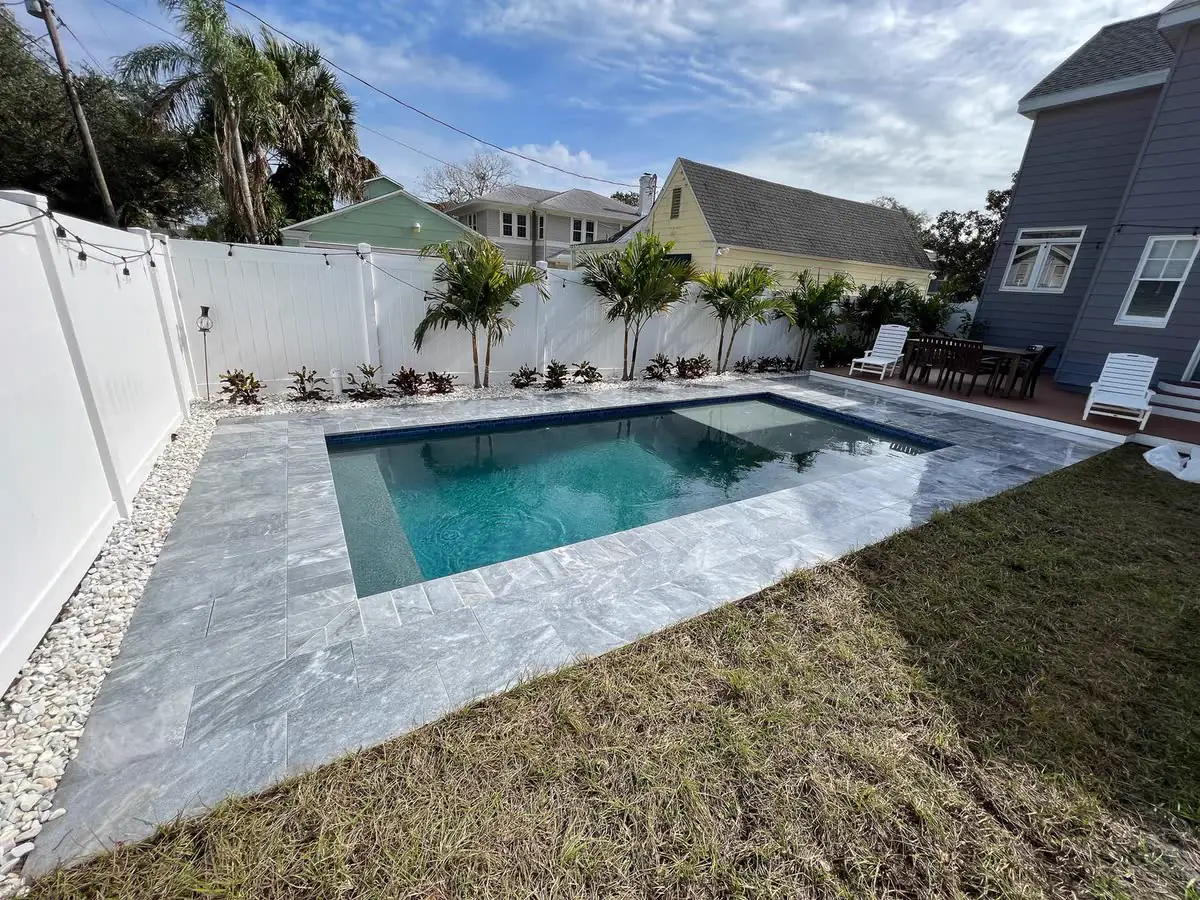 A pale stone pool deck around a rectangular pool and a white fence.