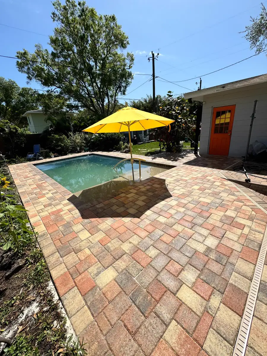 A paver patio in red, tan and grey around a small pool, a yellow umbrella beyond.
