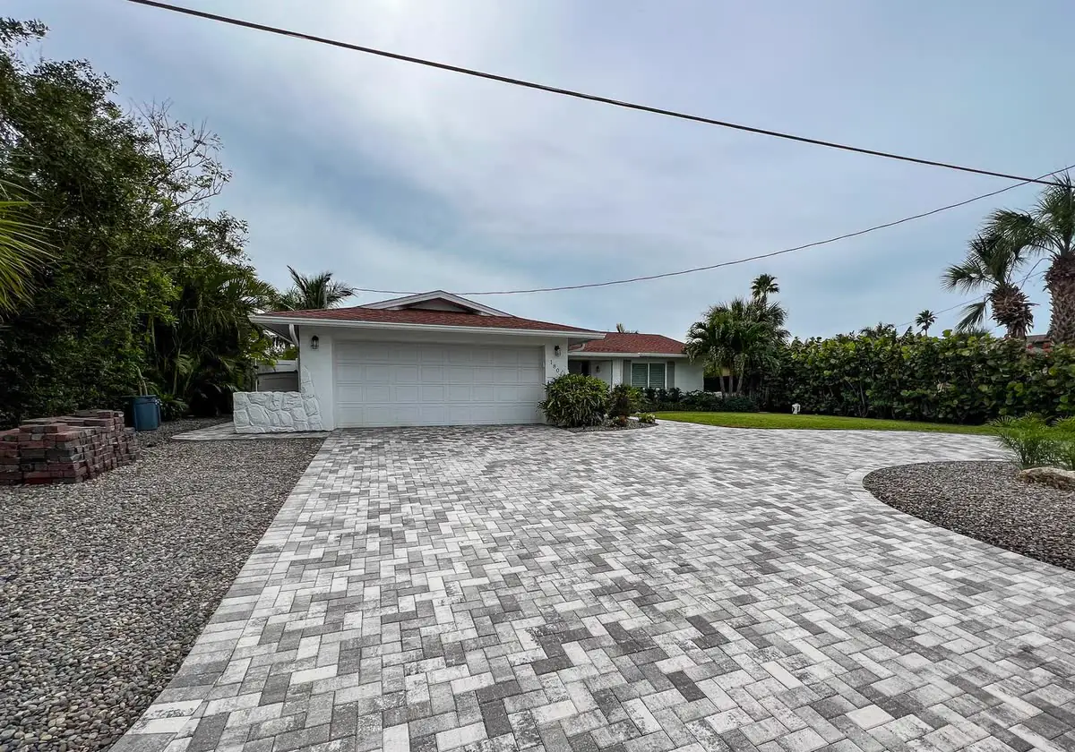 A grey paver driveway beside a gravel yard and a white house.
