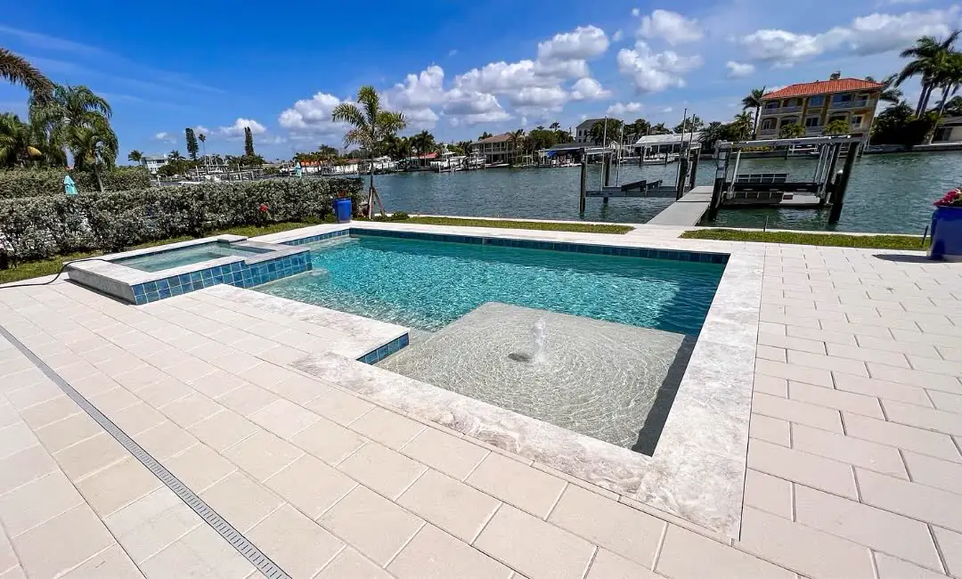 A pale stone pool deck and spa on a canal, boats and docks across the water.