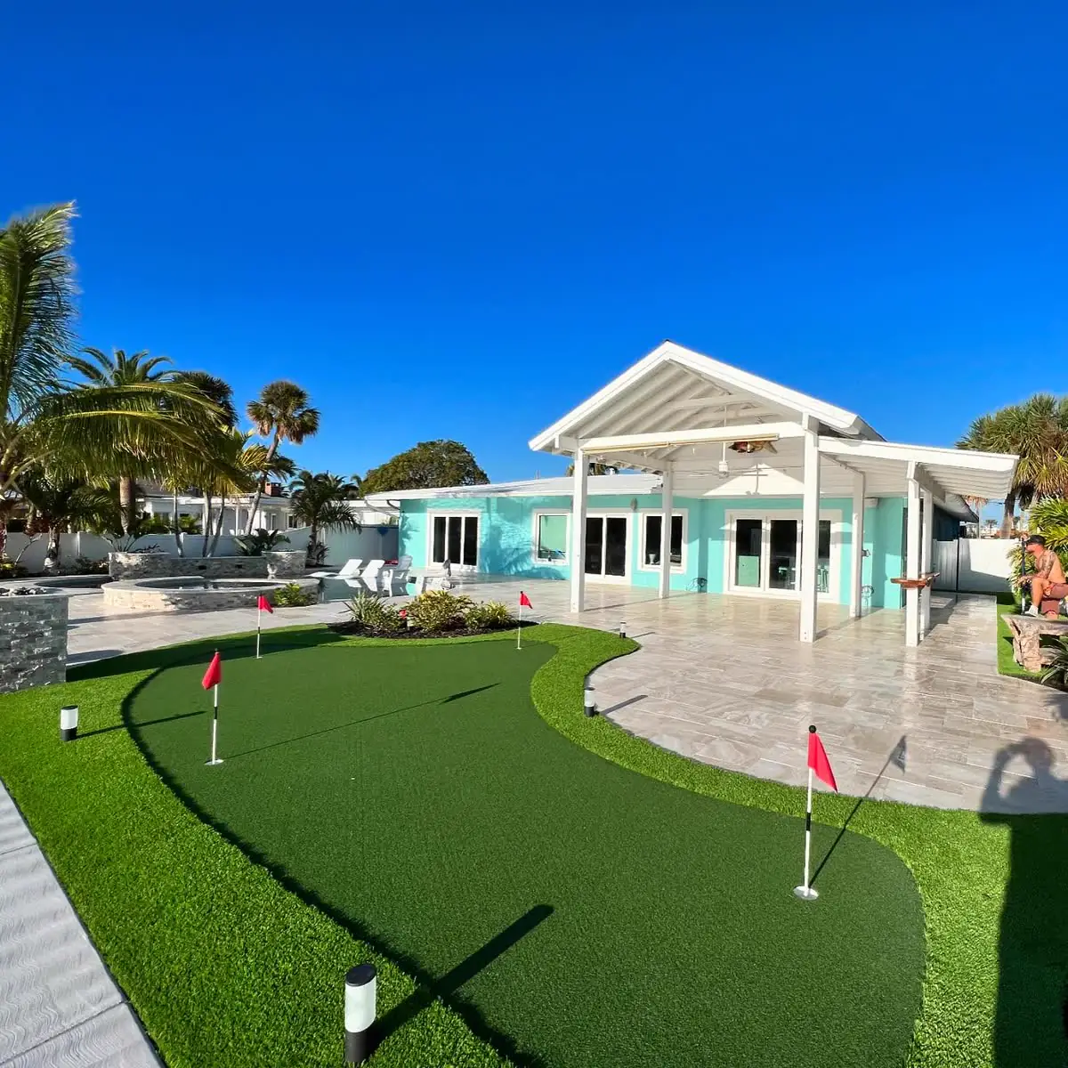 A travertine patio beside a putting green behind a mint-green house.