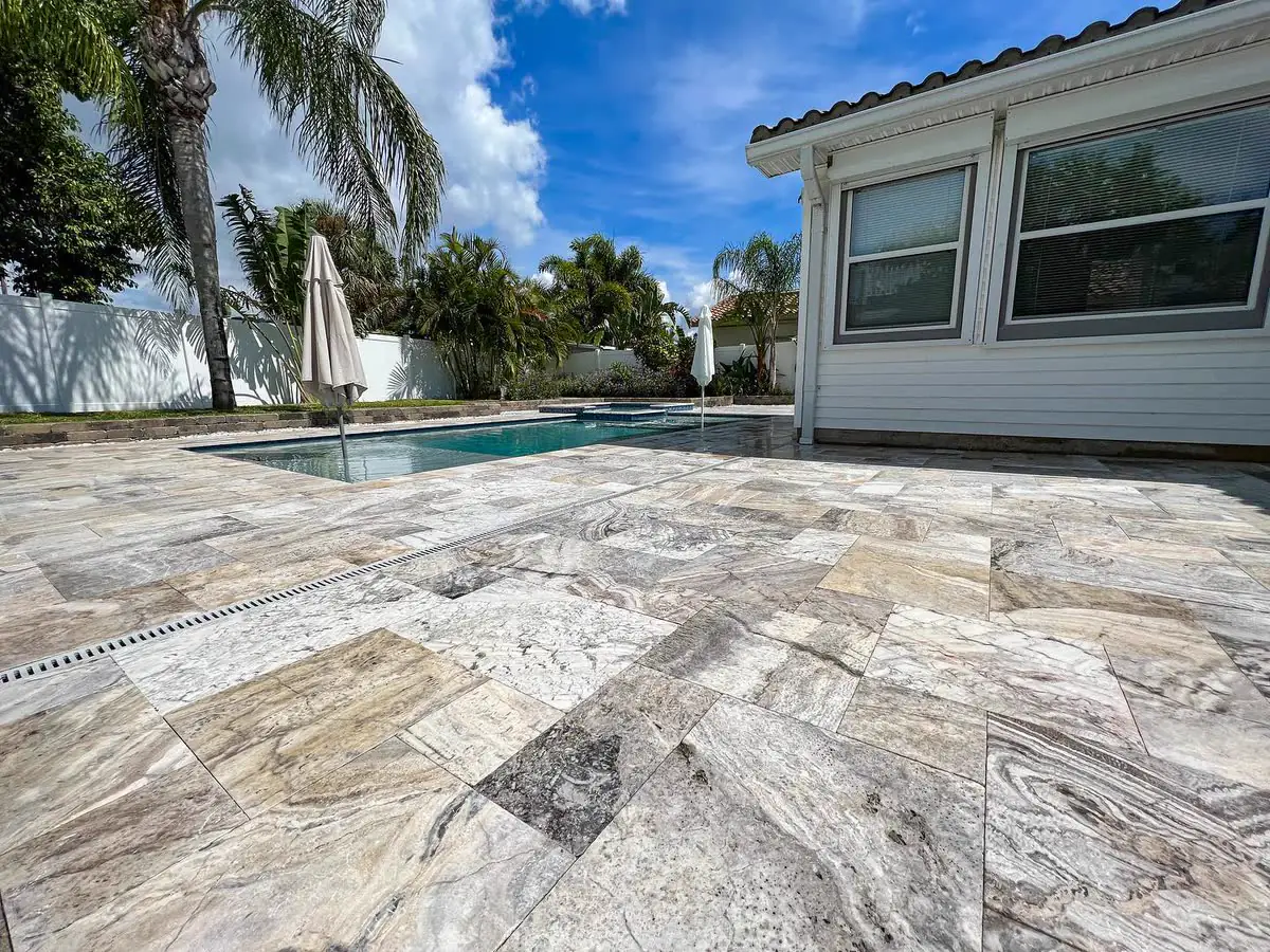 A sealed travertine pool deck beside a white house and palms.